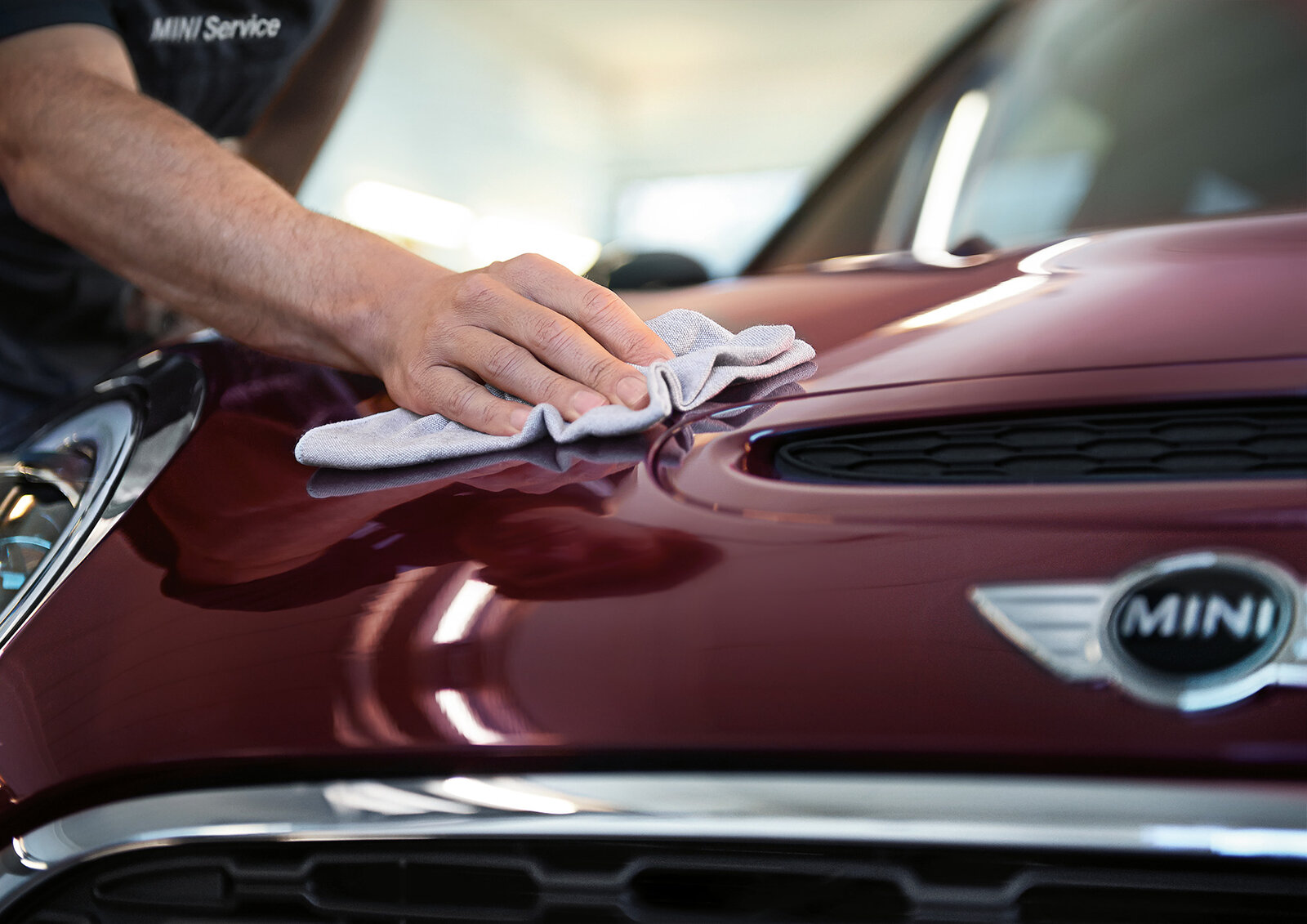 Ein Mitarbeiter eines MINI-Services reinigt die Motorhaube eines roten Autos mit einem Mikrofasertuch. Die Oberfläche glänzt, und der Fokus liegt auf der sorgfältigen Pflege des Fahrzeugs.