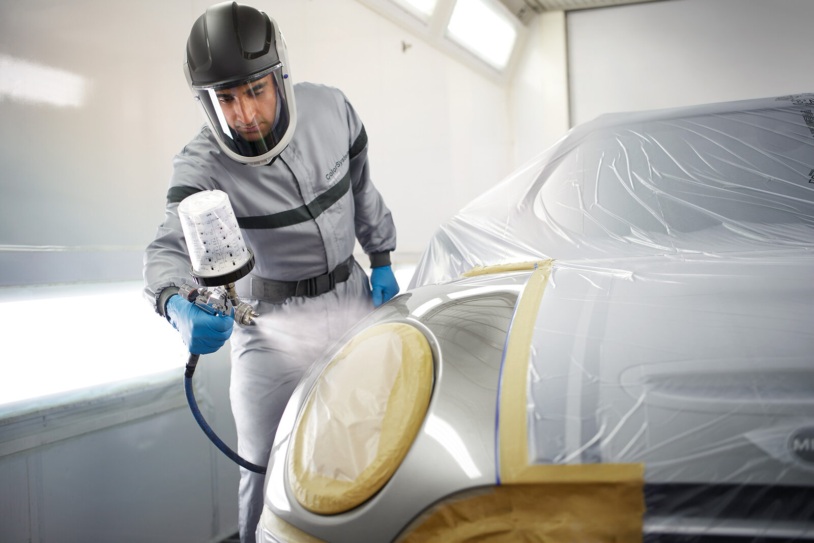 Ein Mann in Schutzkleidung sprüht Farbe mithilfe eines Sprühgeräts auf ein Auto. Der Vorderteil des Fahrzeugs ist teilweise mit Folie abgedeckt, um unerwünschte Farbspritzer zu vermeiden. Der Hintergrund zeigt eine helle, saubere Umgebung.