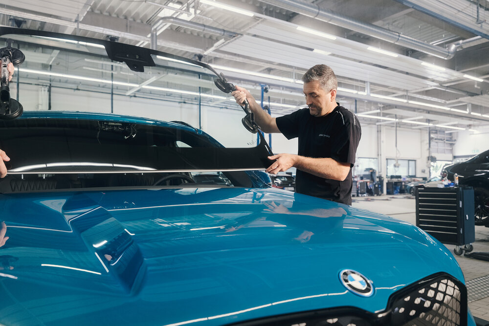 Ein Mechaniker befestigt eine Windschutzscheibe an einem blauen BMW in einer Werkstatt. Er verwendet Saugnäpfe, um die Scheibe präzise zu positionieren. Werkzeug und andere Fahrzeuge sind im Hintergrund sichtbar.