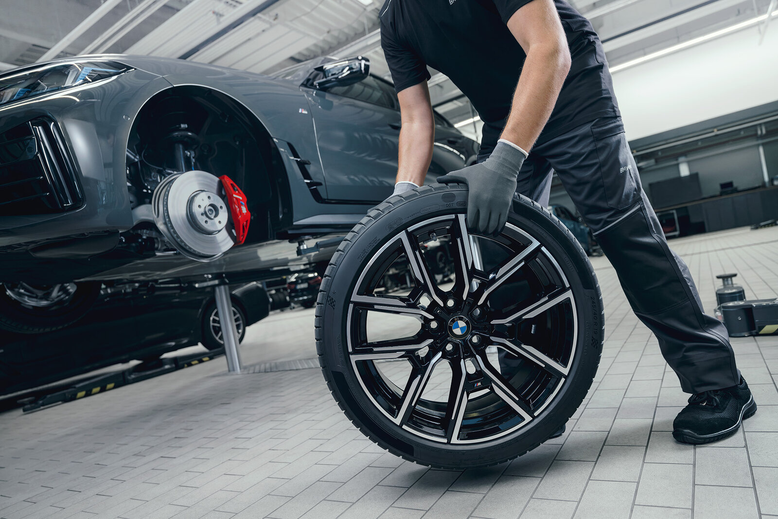 Ein Mechaniker hebt einen neuen Reifen mit einem charakteristischen BMW-Logo an. Der Wagen steht auf einer Hebebühne, während das andere Rad teilweise sichtbar ist. Die Werkstatt hat einen modernen, gut beleuchteten Eindruck. Der Mechaniker trägt Handschuhe und schwarze Arbeitskleidung.