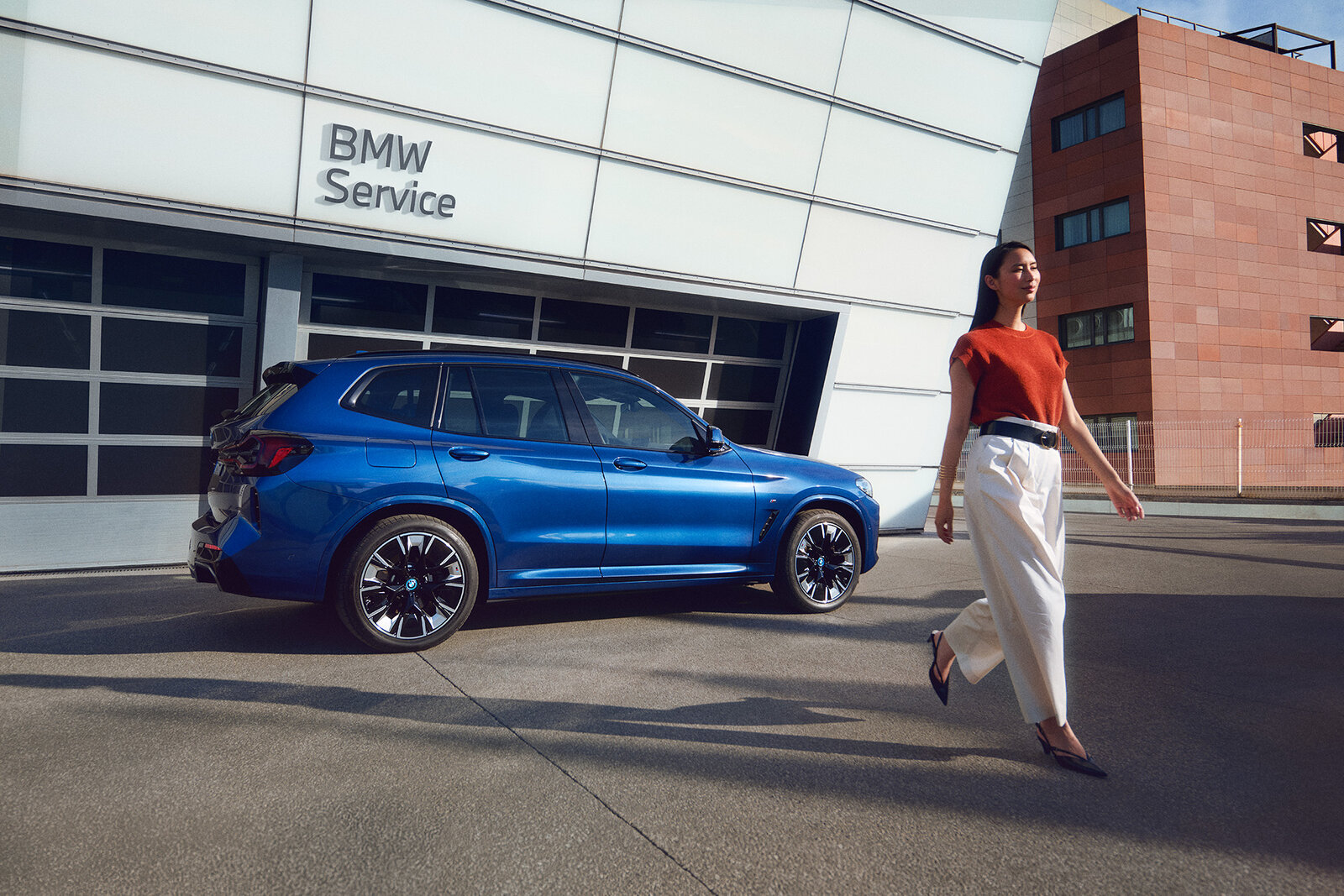 Eine Frau in einem roten Oberteil und weißen Hosen geht an einem blauen BMW SUV vorbei, der vor einem modernen Gebäude mit der Aufschrift BMW Service parkt. Die Szene vermittelt ein Gefühl von Eleganz und urbanem Lifestyle.