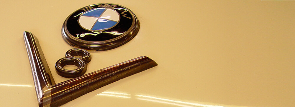 Ein close-up der Motorhaube eines Autos in einer beigen Farbe. Oben befindet sich das BMW-Logo mit einem blauen und weißen Emblem. Daneben sind die Modellnummer und ein charakteristischer Chromstreifen zu sehen.