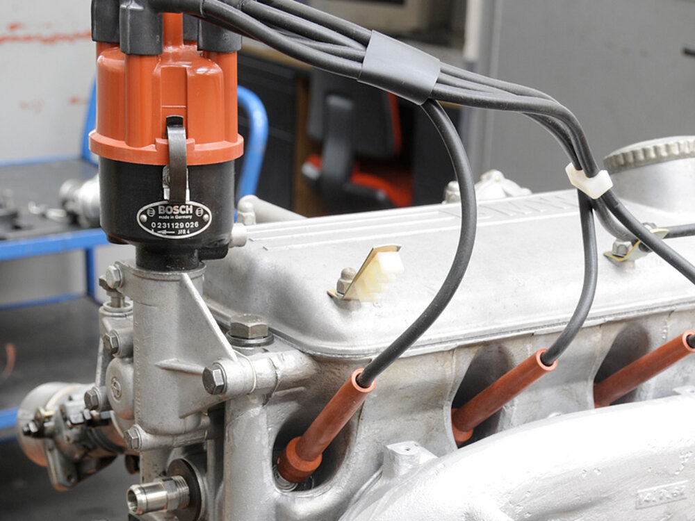 Nahaufnahme eines Motorblocks mit Zündspule und Zündkabeln. Die Zündspule ist rot und trägt das Bosch-Logo. Der Motorblock ist aus silberfarbenem Metall und zeigt Details wie Anschlussstücken und Schraubenelementen. Der Hintergrund ist verschwommen, was den Motor in den Fokus rückt.