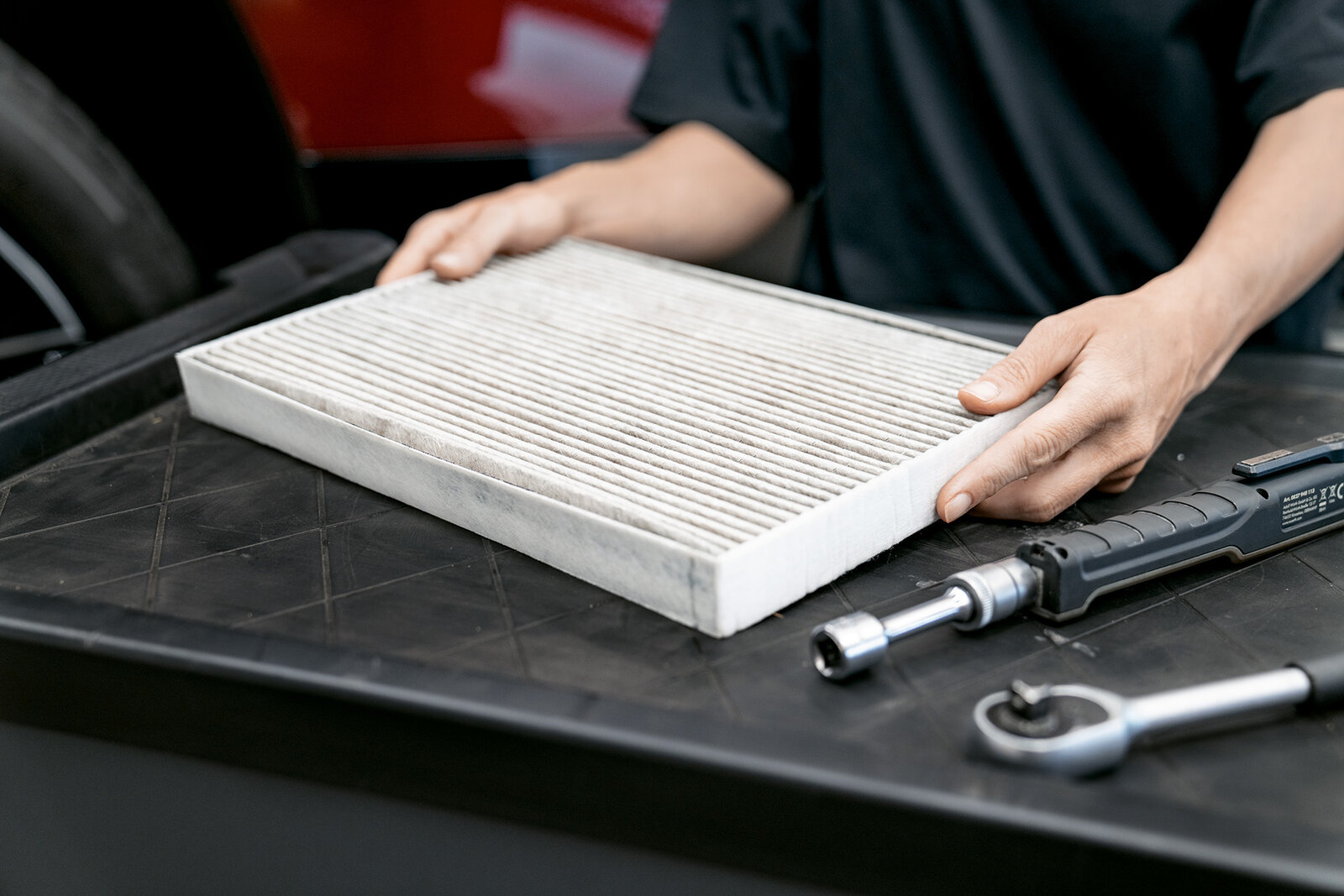 Eine Person hält einen Autofilter, der auf einem schwarzen Werkstatttisch liegt. Neben dem Filter liegen eine Ratsche und ein weiteres Werkzeug. Der Hintergrund ist unscharf und zeigt Teile eines Fahrzeugs.