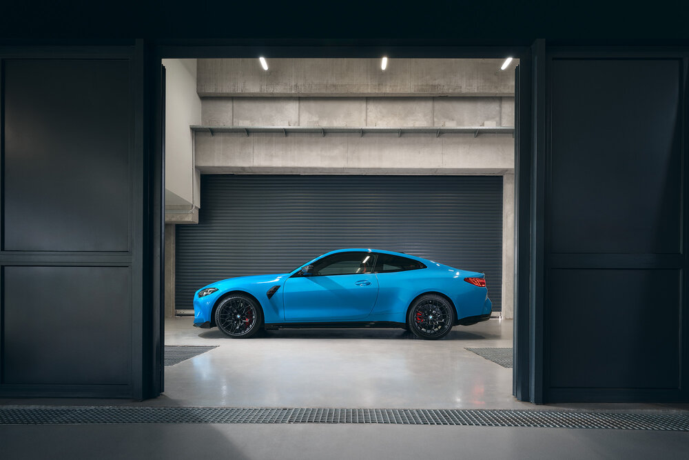 Ein blauer Sportwagen steht in einer modernen, minimalistischen Garage. Er wird seitlich betrachtet und hebt sich durch seine Farben und Formen hervor. Die Wände sind aus Beton, und eine große Rolltür ist im Hintergrund geschlossen.