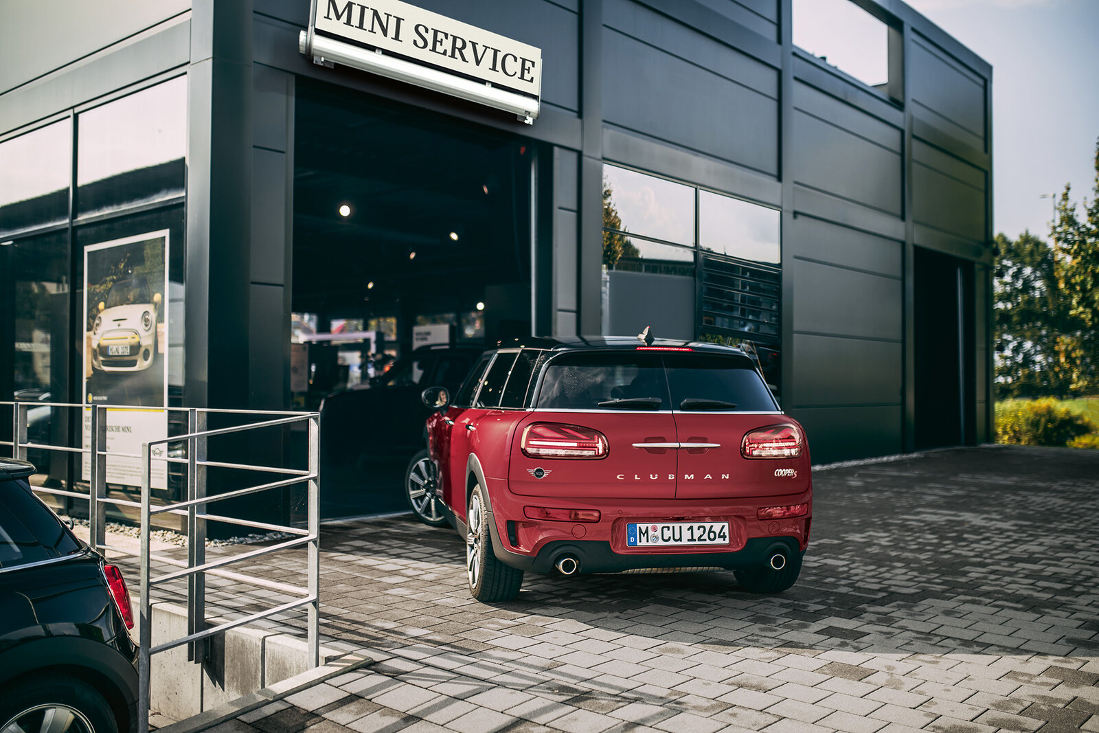 Ein roter Mini Clubman steht vor einem modernen Servicegebäude mit der Aufschrift "MINI SERVICE". Die Fassade ist aus dunklem Material und große Fenster ermöglichen einen Blick auf weitere Autos im Inneren. Die Umgebung wirkt hell und freundlich.