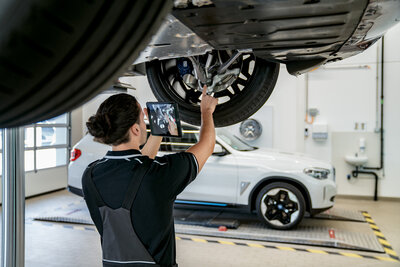 Ein Mechaniker steht unter einem Auto und hält ein Tablet, um Details am Fahrwerk zu überprüfen. Im Hintergrund ist ein weiteres Auto sichtbar. Die Werkstatt hat helle Wände und eine gut beleuchtete Arbeitsumgebung.