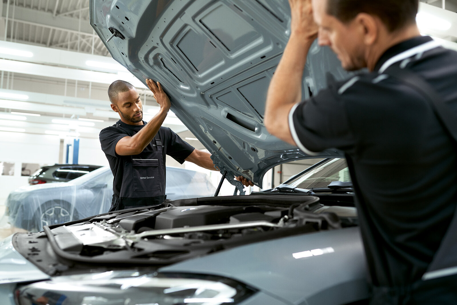 Zwei Mechaniker arbeiten in einer Autowerkstatt. Einer hebt die Motorhaube eines Fahrzeugs an, während der andere dabei hilft. Beide tragen schwarze Arbeitskleidung mit dem Aufdruck BMW Service. Im Hintergrund sind weitere Fahrzeuge sichtbar.