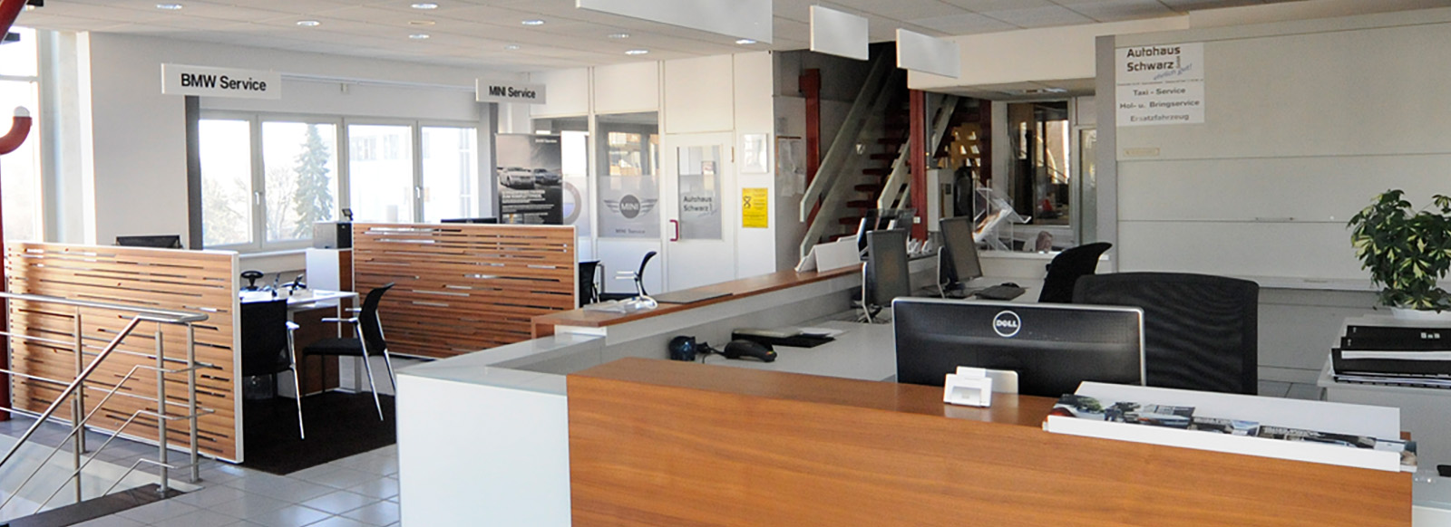 Ein heller Empfangsbereich eines Autohauses mit Schaltern für BMW- und MINI-Service. Am Empfangstresen stehen Computer. Im Hintergrund sind Fenster und Stühle zu sehen. Rechts befindet sich eine Pflanze. Alles wirkt modern und einladend.