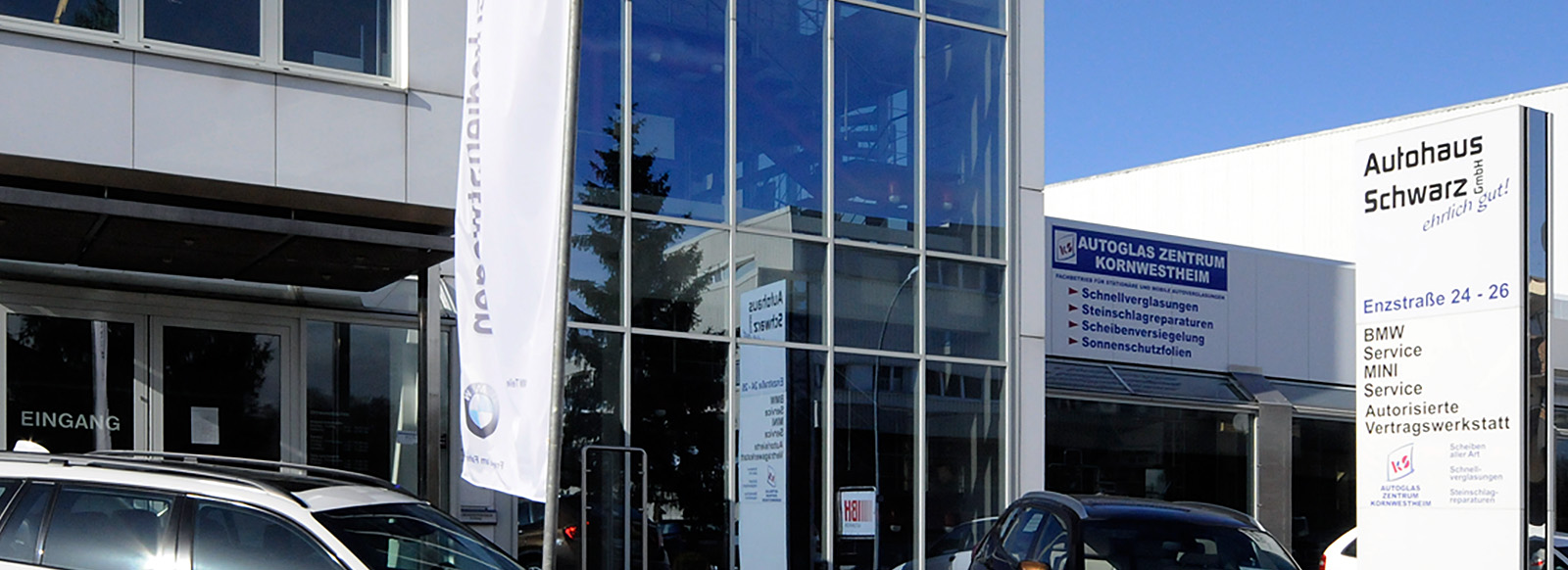 Eingang zum Autohaus Schwarz, einem Autorisierter BMW- und MINI-Service in Kornwestheim. Im Vordergrund stehen Fahrzeuge, während an der Fassade verschiedene Dienstleistungen angezeigt werden, darunter Autoglas-Reparaturen. Der Himmel ist blau und es ist sonnig.