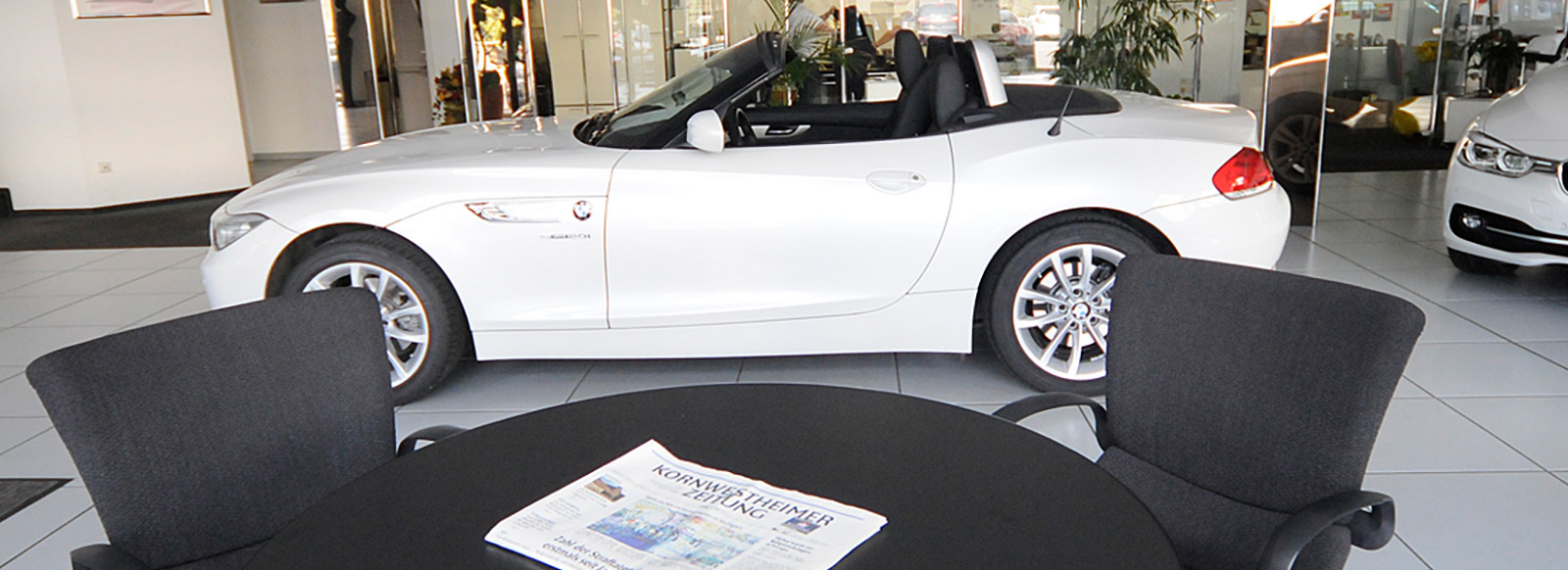 Im Bild ist ein weißes Cabrio zu sehen, das in einem Autohaus steht. Vor dem Fahrzeug befindet sich ein runder Tisch mit einer aufgeschlagenen Zeitung. Der Innenraum des Autohauses ist hell und modern gestaltet, mit einigen Pflanzen im Hintergrund.