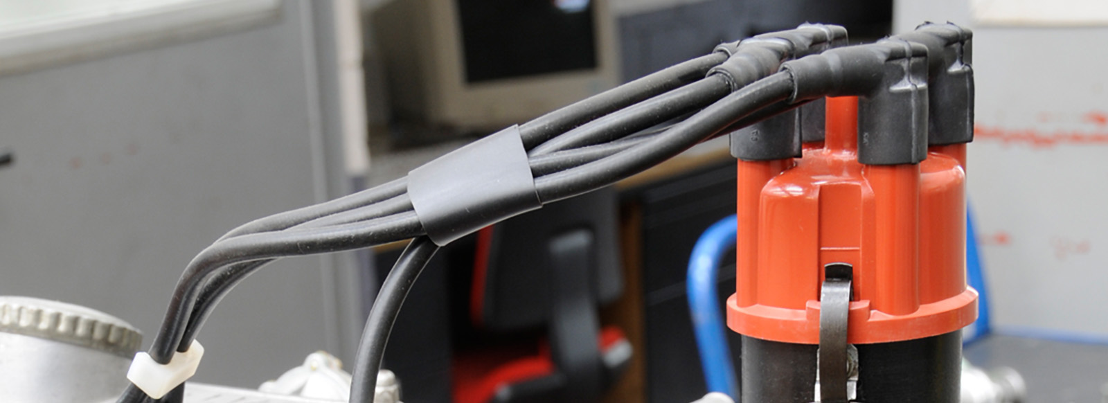 Eine schwarze Zündspule mit mehreren Kabeln, die aus einer roten Kappe herausführen. Die Kabel sind an ein anderes Teil angeschlossen, das nicht genau sichtbar ist. Der Hintergrund zeigt verschwommenen Labor- oder Werkstattraum.
