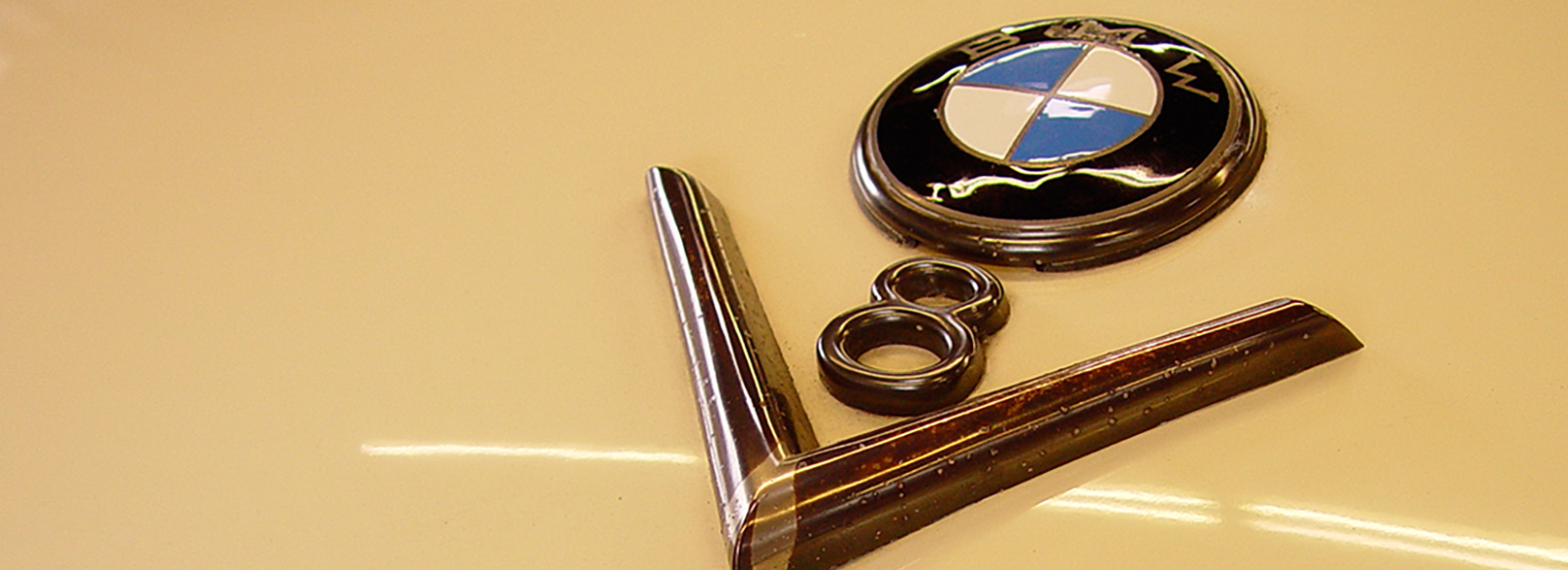 Das Bild zeigt das Emblem einer BMW-Marke, das oval und in den Farben Blau, Weiß und Schwarz gestaltet ist. Daneben befindet sich eine verchromte Zierleiste, die die Zahl 8 darstellt, was auf einen V8-Motor hinweist. Der Hintergrund ist einheitlich beige.